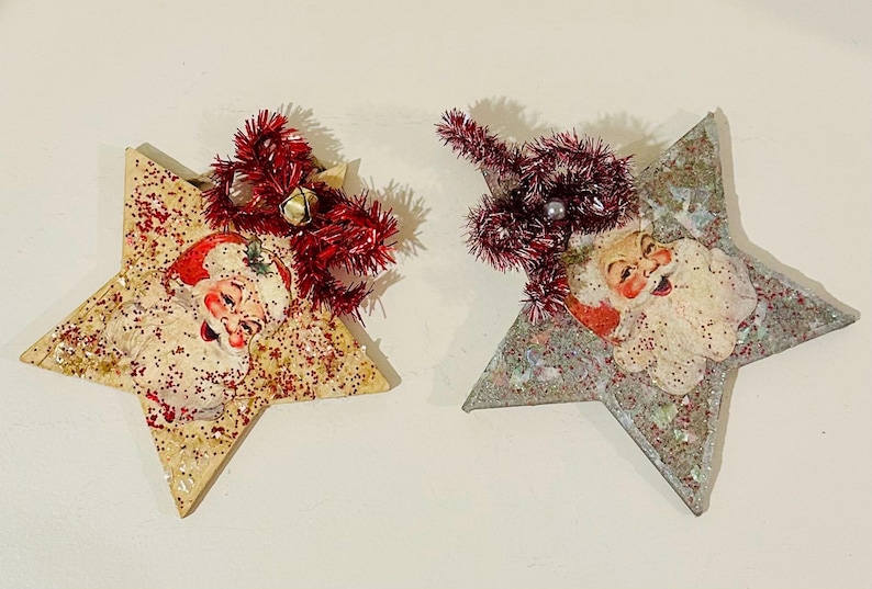 May include: Two star-shaped ornaments featuring vintage-style Santa Claus illustrations. The left star is cream with red glitter, while the right star is silver with iridescent glitter. Both are topped with red tinsel bows and small bells.