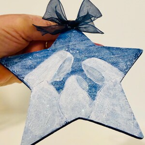 May include: A star-shaped ornament painted in deep blue with a white silhouette of a nativity scene featuring three figures. The surface has a subtle glitter finish and is topped with a dark blue sheer organza ribbon tied in a small bow.