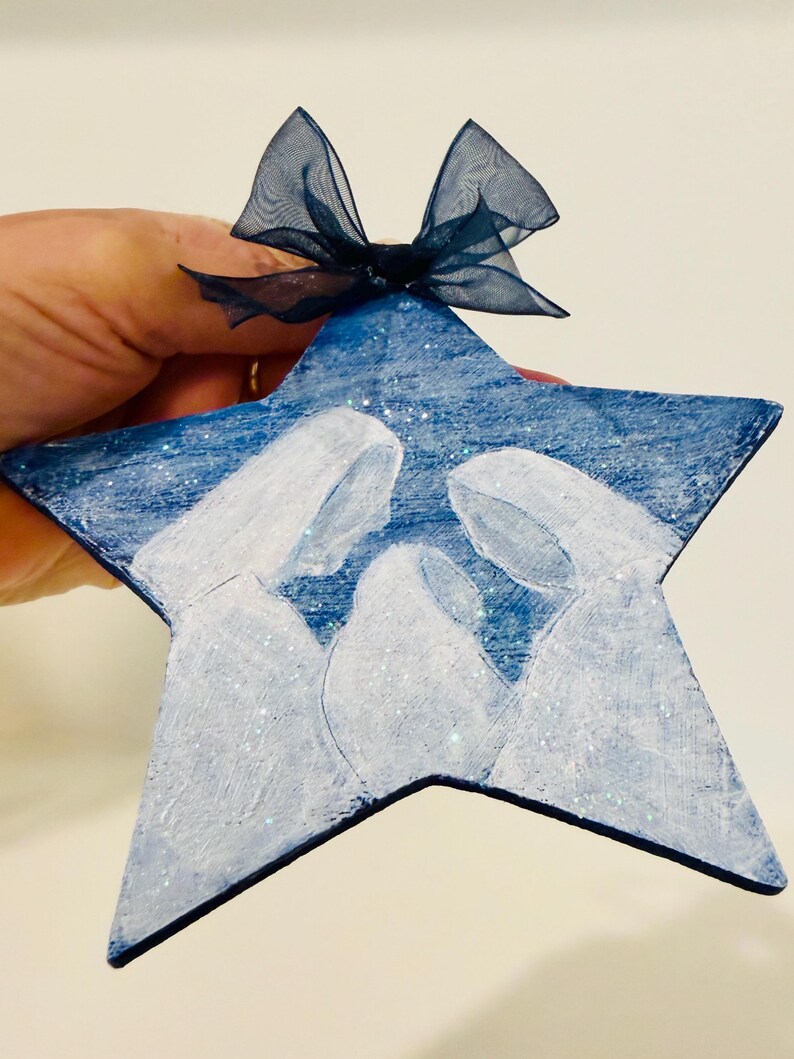 May include: A star-shaped ornament painted in deep blue with a white silhouette of a nativity scene featuring three figures. The surface has a subtle glitter finish and is topped with a dark blue sheer organza ribbon tied in a small bow.