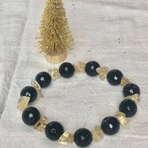 May include: A beaded bracelet featuring alternating large, round, faceted black beads and smaller, irregular, translucent yellow-gold crystal chips. The bracelet is arranged in a circular shape alongside a small, gold-colored decorative tree.