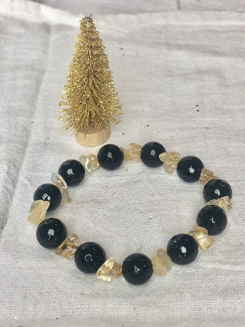 May include: A beaded bracelet featuring alternating large, round, faceted black beads and smaller, irregular, translucent yellow-gold crystal chips. The bracelet is arranged in a circular shape alongside a small, gold-colored decorative tree.