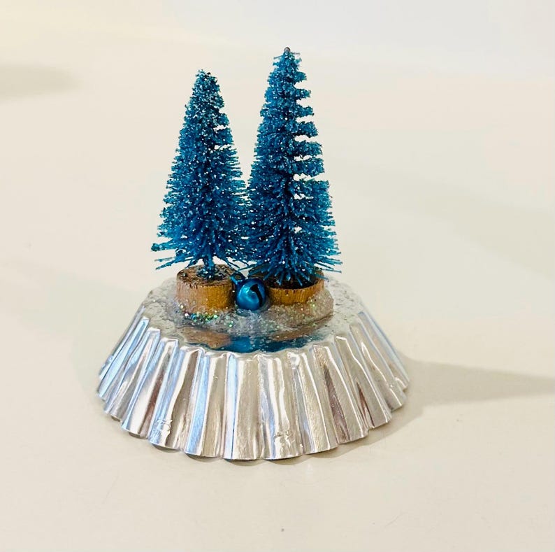 May include: Two miniature blue bottle brush trees with glittery textures stand on a silver fluted metal base. The trees are anchored in small brown wooden bases, and a single small blue jingle bell is placed between them on the snowy surface.
