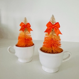 May include: Two miniature bottle brush trees with an ombre color gradient transitioning from cream at the top to bright orange at the base. Each tree is topped with a vibrant orange satin ribbon bow and is set inside a small white ceramic cup.