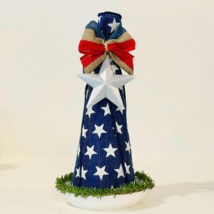 May include: A cone-shaped decorative object covered in blue fabric with a white star pattern. It features a silver star ornament at the center, a red, white, and blue fabric bow at the top, and a ring of green artificial pine needles at the base.