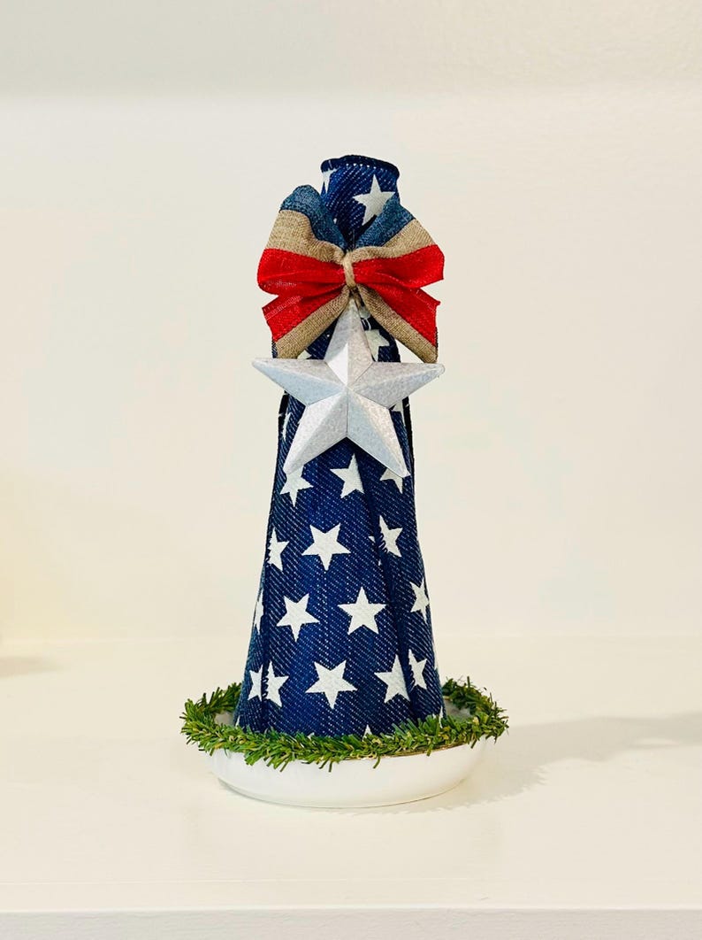May include: A cone-shaped decorative object covered in blue fabric with a white star pattern. It features a silver star ornament at the center, a red, white, and blue fabric bow at the top, and a ring of green artificial pine needles at the base.