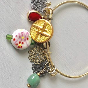 May include: A gold-tone bangle bracelet adorned with various charms. The charms include a red gemstone, a gold button with a star design, a white and pink floral bead, and green stone. Decorative metal filigree elements add detail.