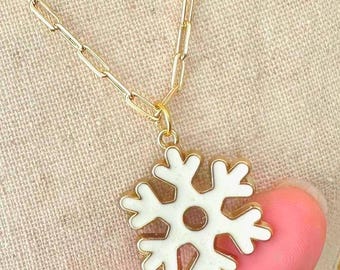 May include: A gold-toned necklace featuring a paperclip chain and a white enamel snowflake pendant. The pendant has a central circular cutout and six symmetrical arms with branching ends, suspended from a small gold-toned jump ring.