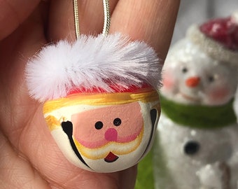 May include: A round metal jingle bell ornament painted to resemble a Santa Claus face with a pink nose, black eyes, and a white beard. The top of the bell is adorned with a fluffy white faux fur trim and features a gold-colored hanging cord.