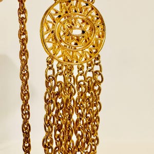 May include: A gold-tone necklace featuring a long, thick twisted chain and a central circular pendant with a radial sunburst pattern. Five shorter chain strands hang from the bottom of the pendant, each ending in a small, round, textured gold-tone disc.