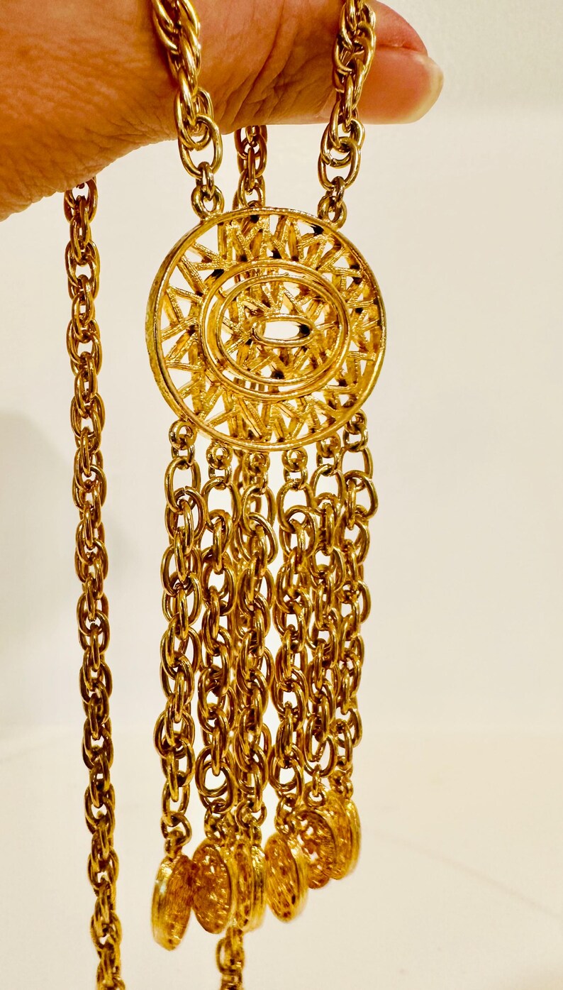 May include: A gold-tone necklace featuring a long, thick twisted chain and a central circular pendant with a radial sunburst pattern. Five shorter chain strands hang from the bottom of the pendant, each ending in a small, round, textured gold-tone disc.