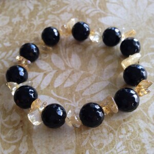 May include: A beaded bracelet featuring alternating large, round, faceted black beads and smaller, irregular, translucent yellow-gold crystal chips arranged in a circular pattern to form a continuous loop of contrasting dark and light elements.