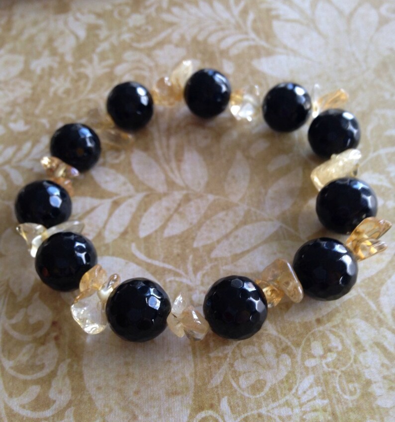 May include: A beaded bracelet featuring alternating large, round, faceted black beads and smaller, irregular, translucent yellow-gold crystal chips arranged in a circular pattern to form a continuous loop of contrasting dark and light elements.