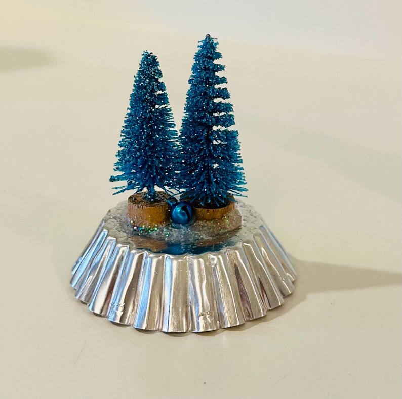 May include: A decorative ornament featuring two miniature blue glittery bottle brush trees mounted on a silver-colored fluted metal tart mold base. The trees are set on a textured, snowy-looking surface with a small blue jingle bell at the base.