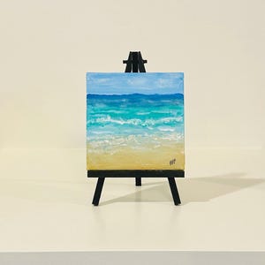 May include: A square canvas painting of a beach scene featuring a sandy yellow shore, turquoise and blue ocean waves, and a light blue sky, displayed on a black wooden tripod easel. The artist's initials BKF are signed in the bottom right corner.