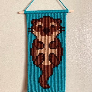 May include: A turquoise and brown string art wall hanging featuring a cartoon otter. The otter is brown with a tan face and has a happy expression. The wall hanging has a wooden dowel at the top and is finished with fringe at the bottom.