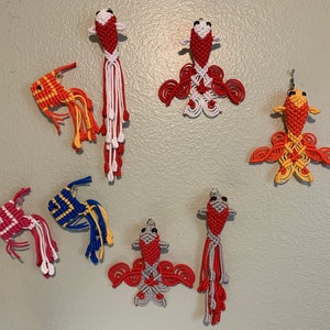 May include: Seven handmade macrame goldfish ornaments in various colors, including red, white, yellow, blue, and gray. The ornaments are hanging on a light gray wall.