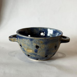 Può includere: Scolapasta in ceramica blu con numerosi fori di drenaggio. La ciotola ha due manici ad anello e una superficie strutturata. Questo scolapasta fatto a mano è progettato per scolare e risciacquare gli alimenti.