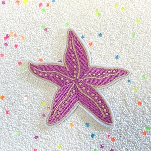 以下が含まれることがあります： ピンクとゴールドの刺繍が施されたヒトデのパッチ。パッチは、カラフルなグリッターが散りばめられた白い背景の上にあります。