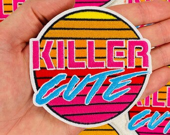 May include: A round, embroidered patch with a sunset design in shades of orange, pink, and yellow. The text "Killer Cute" is embroidered in pink and blue.