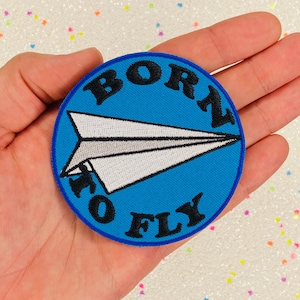 May include: A blue and white embroidered patch with a paper airplane and the text "Born to Fly".