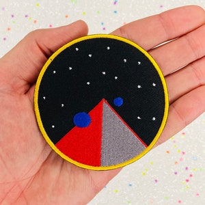 May include: A black, red, and gray embroidered patch with a yellow border. The patch features a stylized mountain with two blue circles above it. The background is black with white stars.