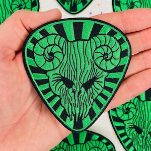 May include: Green and black embroidered iron-on patch with a skull with horns in a sunburst design.