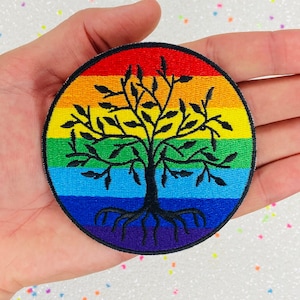 以下が含まれることがあります： 中央に黒い木のシルエットが描かれた、虹色の丸いパッチ。木は根と枝があり、葉が生えています。虹の色は赤、オレンジ、黄色、緑、青、藍色、紫です。