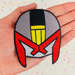 May include: An embroidered patch featuring a helmet with a visor, in red, black, yellow and grey. The helmet is a stylised depiction of a character's head.