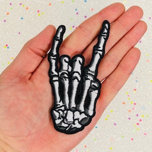 May include: Embroidered patch showing a black and white skeleton hand making the 'rock on' sign. The patch has a black border and detailed stitching. Suitable for application on clothing or accessories.