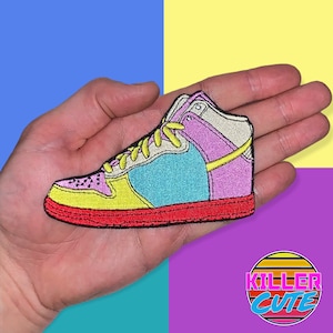May include: A colorful embroidered patch of a high-top sneaker with a yellow, pink, blue, and white color scheme. The patch is held in a hand against a multi-colored background.