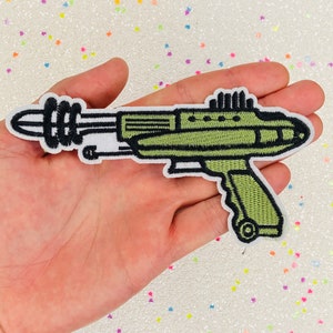 May include: A green and black embroidered iron-on patch of a futuristic ray gun.