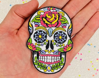 May include: A white sugar skull embroidered patch with colorful flowers, a pink rose, and green leaves.
