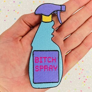 May include: An iron-on patch embroidered in blue and purple, shaped like a spray bottle with the text "BITCH SPRAY" in pink.