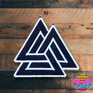 May include: A black and white embroidered patch with a triple triangle design. The patch is on a wooden background.