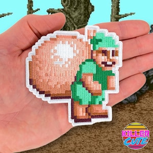 Pode incluir: Um remendo bordado com um personagem pixelado carregando um grande saco nas costas. O personagem está vestindo uma camisa verde e calças marrons. O remendo é cercado por uma borda branca.