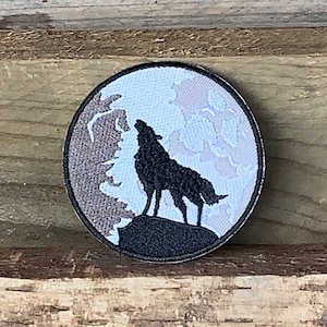 May include: An embroidered patch featuring a black wolf howling at the moon. The moon is a white circle with a grey and white cloud design.