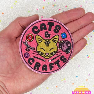 May include: Pink round embroidered patch with the text "Cats & Crafts" in black lettering. A cartoon cat with yellow fur and closed eyes is in the center of the patch. The cat is surrounded by various craft supplies, including scissors, yarn, and buttons.