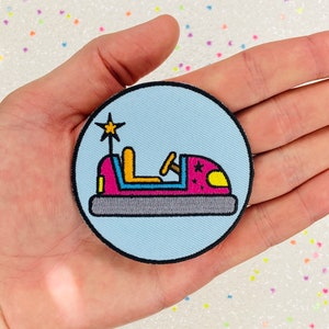 May include: A blue and white embroidered patch of a pink bumper car with a yellow steering wheel and a star on top.
