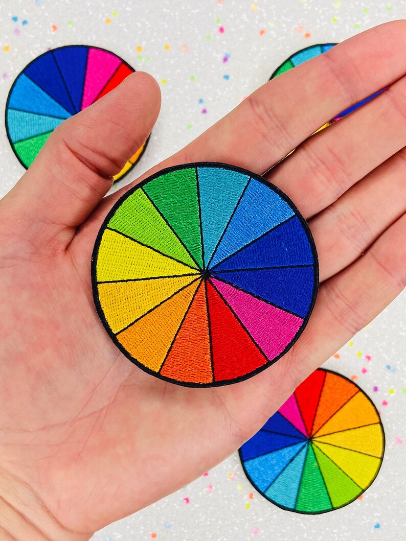 May include: A colorful embroidered patch in the shape of a color wheel. The patch has 12 sections, each a different color of the rainbow.