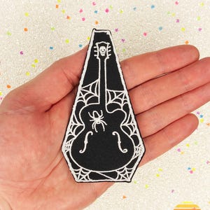 Pode incluir: Etiqueta bordada em preto e branco de uma guitarra com uma aranha e uma caveira na cabeça, cercada por um desenho de teia de aranha.