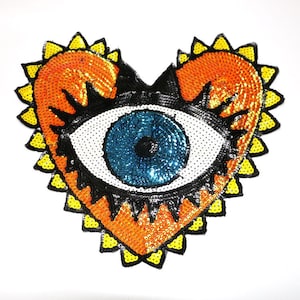 May include: A heart-shaped patch with a large blue eye in the center. The eye is surrounded by white sequins and the heart is outlined in yellow sequins. The heart is orange and black.