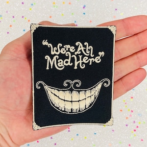 May include: A black embroidered patch with a white outline and the text "We're All Mad Here" and a white grinning Cheshire Cat mouth.