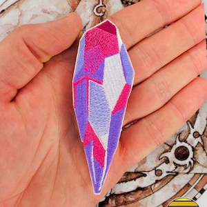 以下が含まれることがあります： 紫、ピンク、白の刺繍が施されたクリスタルパッチ。パッチは長く尖ったクリスタルの形をしています。
