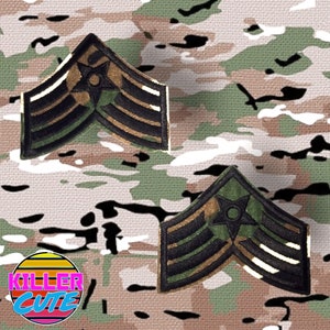 May include: Two black and green military rank insignia patches with gold stripes and a star with a circle in the centre. The patches are on a camouflage background.