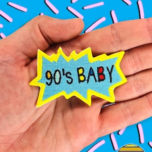 May include: A blue and yellow embroidered patch with the text "90's BABY" in colourful letters. The patch is in the shape of a speech bubble.