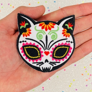 May include: White embroidered iron-on patch of a cat skull with Day of the Dead sugar skull details. The cat has black eyes, a pink nose, and red and yellow flowers on its head.