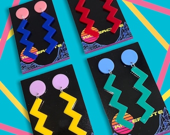 May include: Four pairs of earrings with a zig-zag design. The earrings are on black cards with a retro design and the text "Hi-er-cute". The earrings are yellow, red, blue, and green.