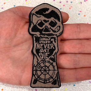 May include: A brown embroidered patch featuring a skull, a compass, and the text "Never Say Die".