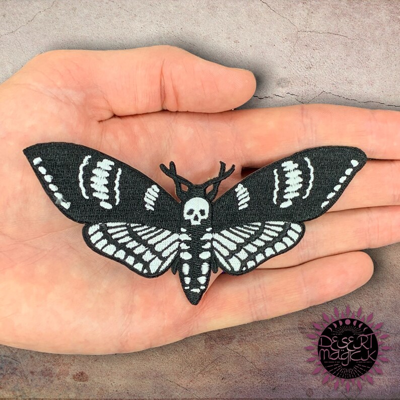 May include: Black and white embroidered iron-on patch of a moth with a skull and antlers. The moth has a detailed design with intricate patterns on its wings.
