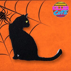Pode incluir: Patch de silhueta de gato preto com olhos amarelos, em um fundo laranja com uma teia de aranha preta. O patch tem o texto "Killer Cute" em uma fonte de estilo retrô.